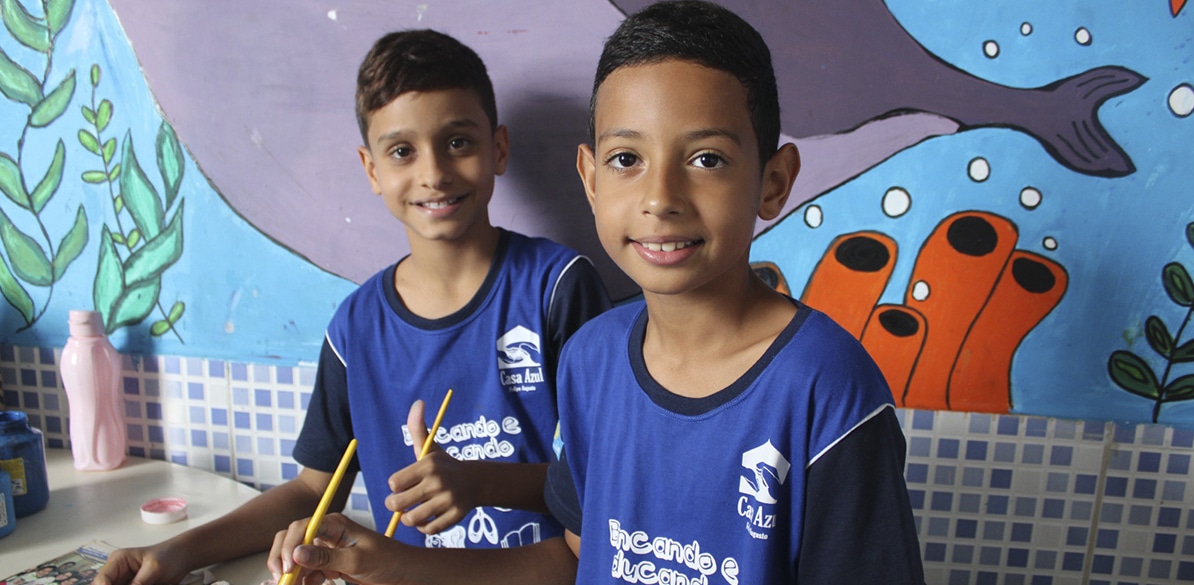 Dois meninos sorrindo com camisetas azuis da Casa Azul, segurando lápis durante atividade educativa, com mural colorido de fundo na instituição em Brasília.
