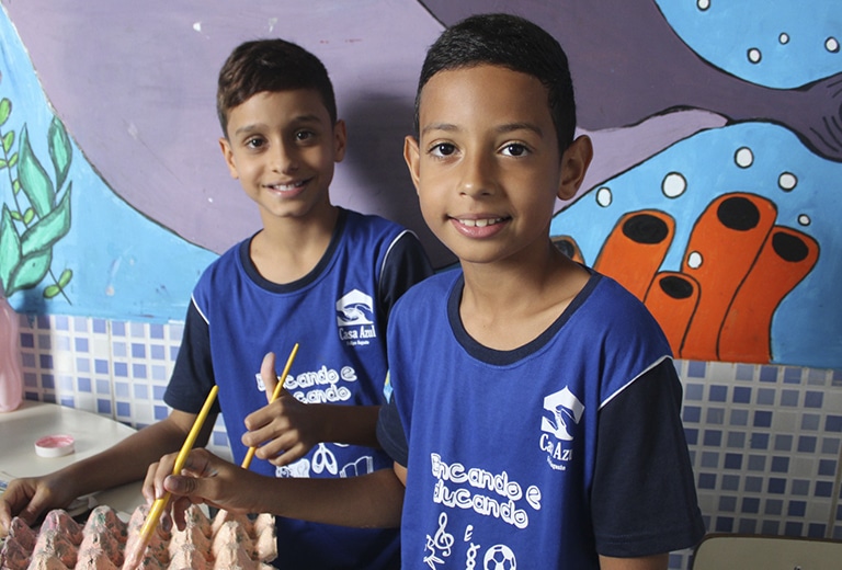 Dois meninos sorrindo com camisetas azuis da Casa Azul, segurando lápis durante atividade educativa, com mural colorido de fundo na instituição em Brasília.