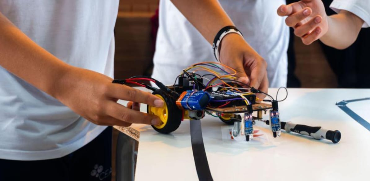 Jovem manipula um veículo robótico de pequeno tamaño sobre uma mesa branca. O robô tem rodas amarelas, sensores frontales e vários cabos conectados a uma placa eletrônica, refletindo os valores do CEAP em Pedreira, SP, em um ambiente educativo prático de tecnologia.