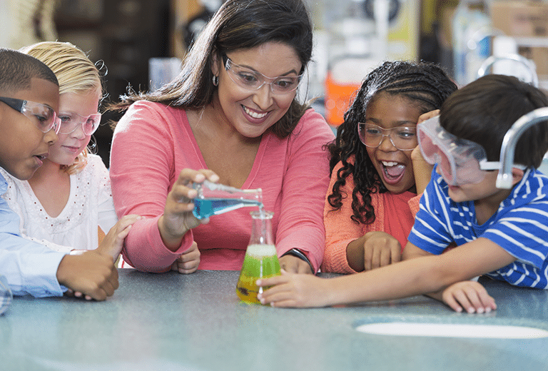 Uma professora orienta um pequeno grupo de crianças enquanto realizam um experimento científico simples. Usando óculos de proteção, elas observam com entusiasmo a mistura de líquidos coloridos em um frasco sobre a mesa, reagindo e mudando de cor.