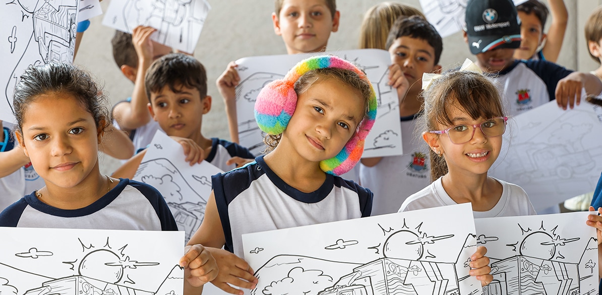 Grupo de crianças participando de uma atividade cultural e educacional em uma sala de aula. Uma delas usa fones de ouvido colorido, outra usa óculos, e todas seguram desenhos para colorir, parte dos materiais e recursos educativos proporcionados pela Fundación MAPFRE.