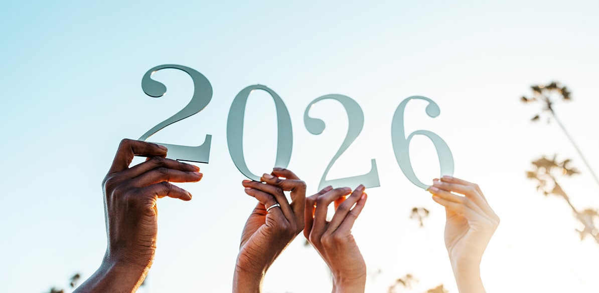 Mãos erguidas em diferentes tons de pele de pessoas segurando grandes números formando 2026 contra um céu claro, simbolizando união e compromisso com o novo ano.