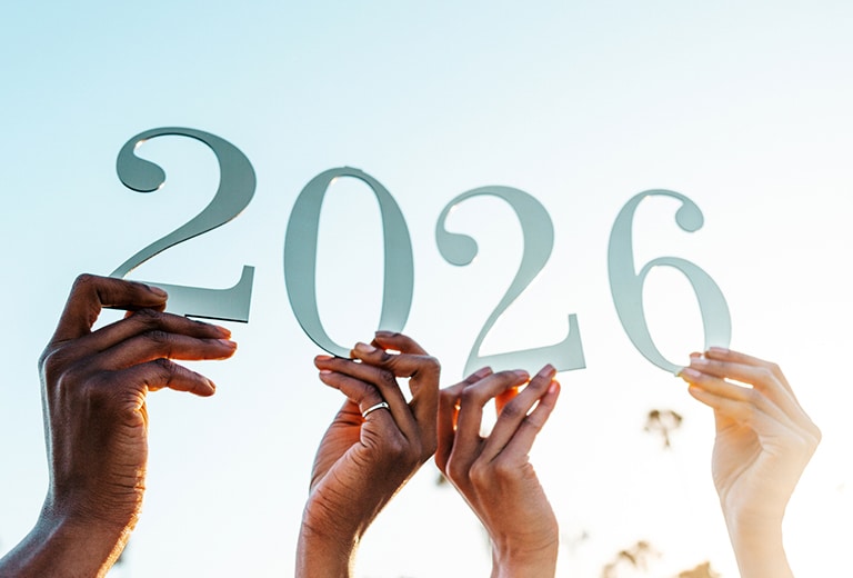 Mãos erguidas em diferentes tons de pele de pessoas segurando grandes números formando 2026 contra um céu claro, simbolizando união e compromisso com o novo ano.