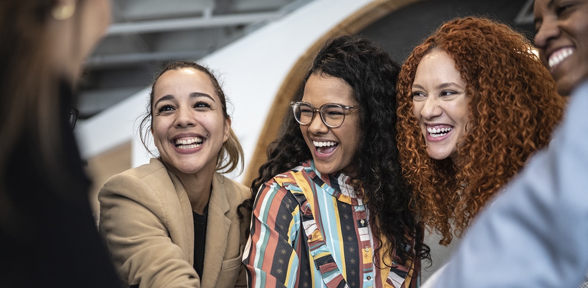 Grupo de mulheres sorrindo e interagindo em um ambiente profissional, representando diversidade, conexão, liderança feminina e celebração do Dia Internacional da Mulher.