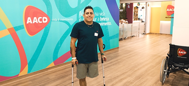 Movimento é vida Homem com muleta caminhando no corredor da AACD em frente a um painel colorido com a marca da instituição.