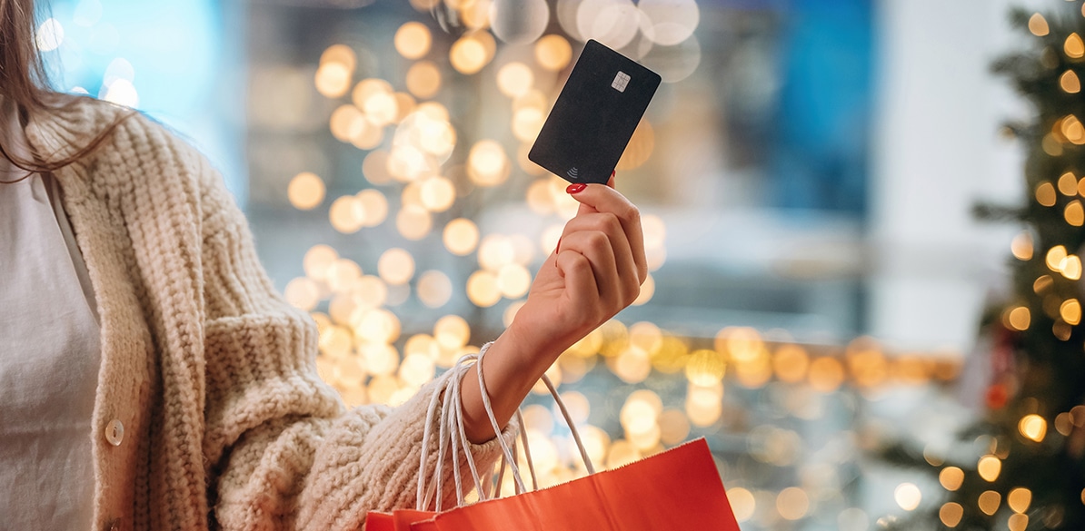 Mão de mulher com unhas vermelhas segurando um cartão de crédito preto acima de uma sacola de compras laranja, com luzes douradas desfocadas e árvore de Natal ao fundo.