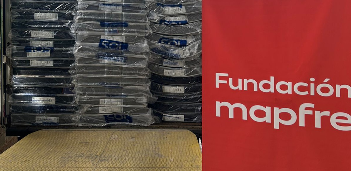 Ajuda humanitária Pallet com várias bandejas plásticas pretas empilhadas e embaladas em plástico transparente, ao lado de um painel vermelho com o texto “Fundación Mapfre”.
