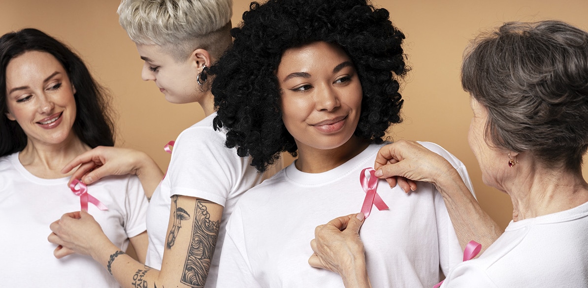 Outubro Rosa no Brasil Imagem de quatro pessoas de diferentes idades e etnias, usando camisetas brancas, unidas por uma causa em apoio ao câncer de mama. Cada uma delas está recebendo um laço rosa em seus peitos. Da esquerda para a direita, a primeira mulher sorri enquanto alguém coloca o laço rosa em sua camiseta. A segunda pessoa é uma mulher com cabelo curto e tatuagens visíveis, que parece estar sorrindo enquanto observa o laço que lhe colocaram. A terceira mulher é de pele morena e cabelos cacheados, sorrindo suavemente enquanto alguém, provavelmente uma mulher idosa, coloca um laço rosa em sua camisa. Ao fundo, a mulher idosa, com cabelos grisalhos e um semblante gentil, está colocando o laço em outra pessoa. A cena transmite uma mensagem de apoio e solidariedade ao câncer de mama, destacando a união e a força feminina.