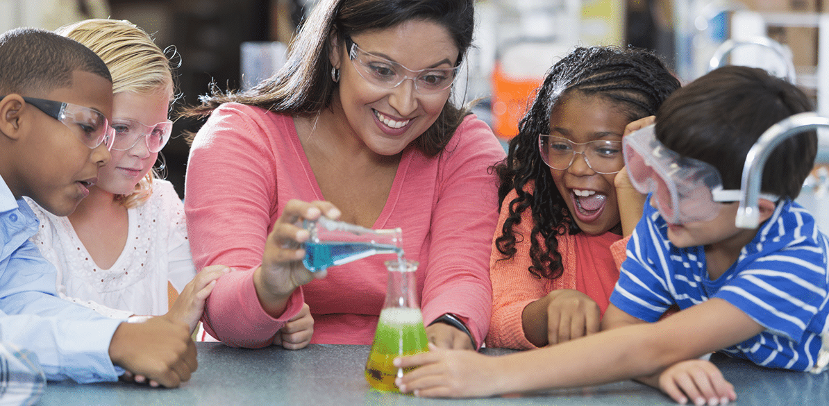 Uma professora orienta um pequeno grupo de crianças enquanto realizam um experimento científico simples. Usando óculos de proteção, elas observam com entusiasmo a mistura de líquidos coloridos em um frasco sobre a mesa, reagindo e mudando de cor.