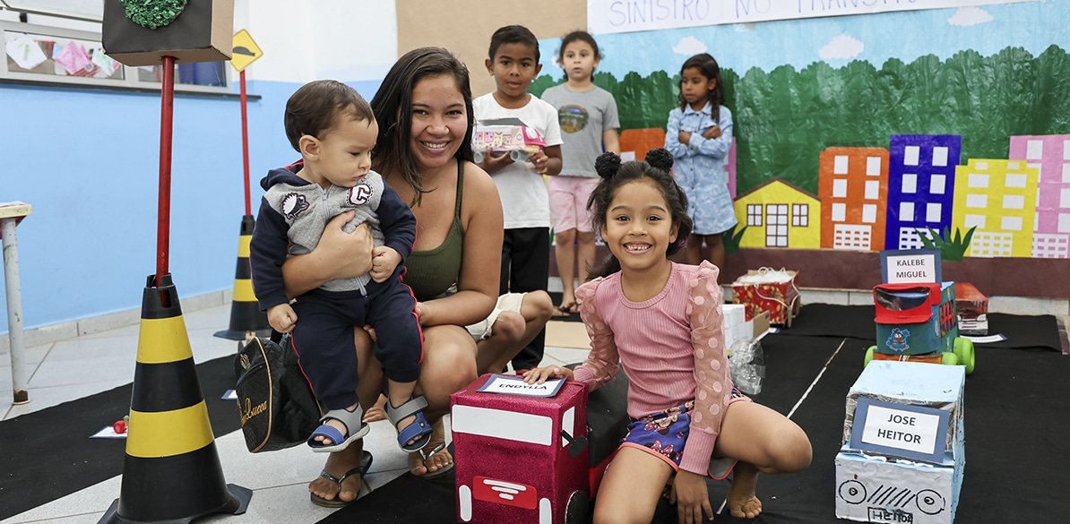 educacion-viaria-1194x585-1 Mãe sorridente segurando um bebê ao lado de uma menina pequena em frente a maquetes de carros e edifícios coloridos, durante atividade educativa sobre mobilidade urbana e segurança viária em sala de aula, com outras crianças ao fundo.