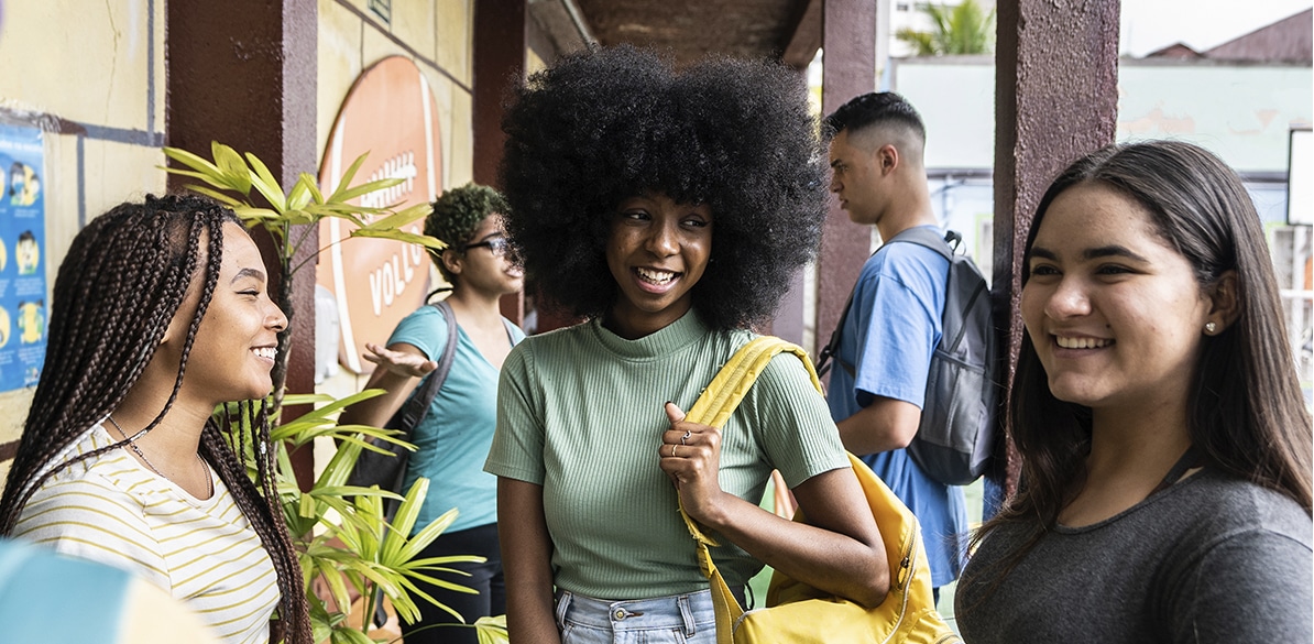 Três jovens em primeiro plano, sorrindo e conversando num espaço externo que parece a entrada de um centro educativo, com plantas ao lado e um corredor coberto ao fundo. A pessoa ao centro carrega uma mochila amarela, enquanto as outras duas se voltam para ela, sugerindo um momento de troca e convivência. Ao fundo, outros jovens circulam pelo local, reforçando a ideia de comunidade e participação. 