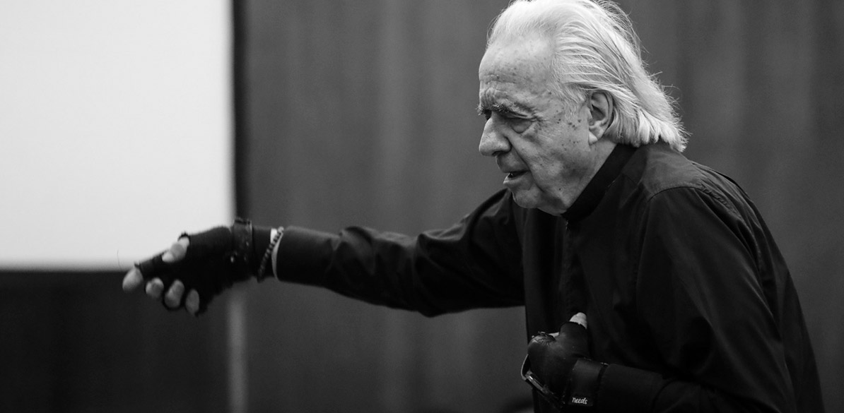 Fotografia em preto e branco do maestro João Carlos Martins de perfil, usando luvas e camisa negras. Ele aparece em um gesto expressivo, regendo a Orquesta Jovem em uma apresentação durante a visita à Fundação Bachiana, em São Paulo, em homenagem à sua trajetória e impacto social.