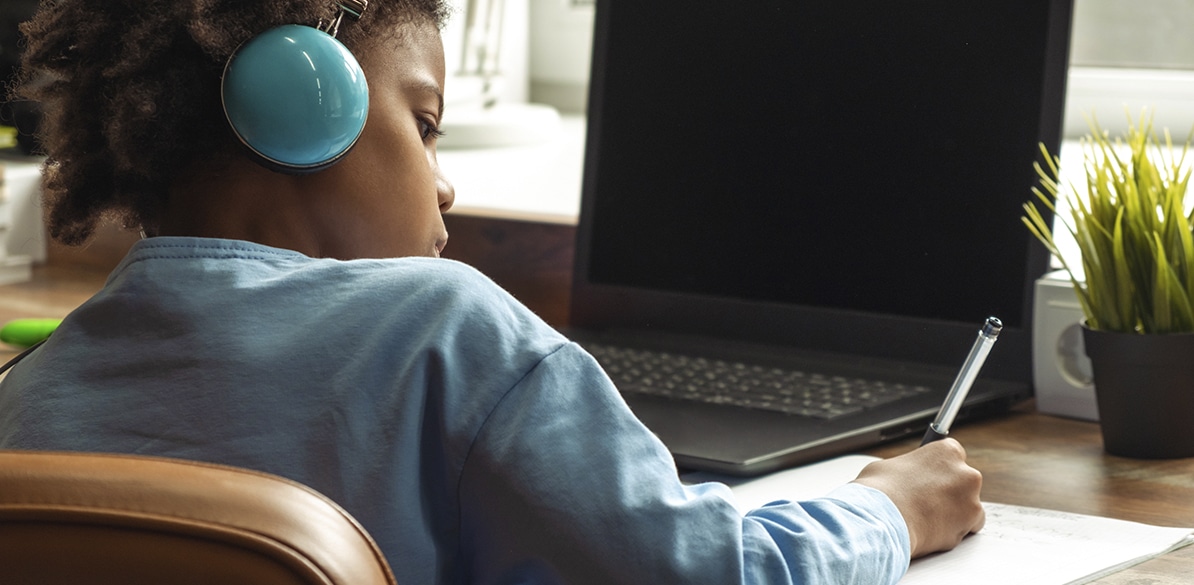 Criança de costas com fones de ouvido azuis escrevendo em um caderno diante de um laptop aberto em uma mesa de madeira, representando o uso da internet por menores em ambiente doméstico.