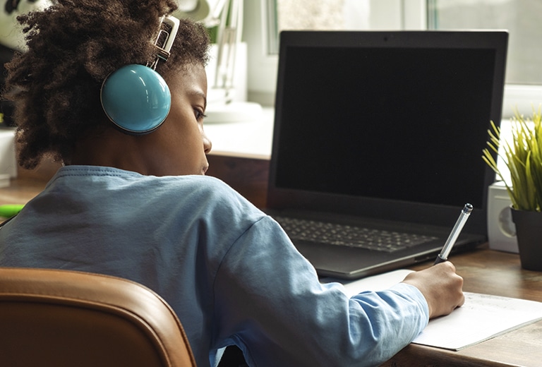 Criança de costas com fones de ouvido azuis escrevendo em um caderno diante de um laptop aberto em uma mesa de madeira, representando o uso da internet por menores em ambiente doméstico.