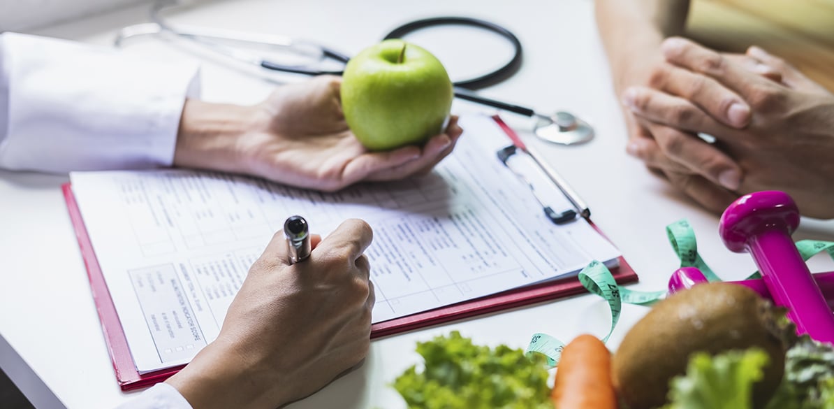 Médico y paciente adultos en consulta: profesional de bata blanca con estetoscopio sostiene una manzana verde saludable sobre mesa con dossier médico, bolígrafo rojo, zanahoria y cinta métrica rosa; fondo blanco resalta hábitos saludables sin dietas rápidas para 2026.