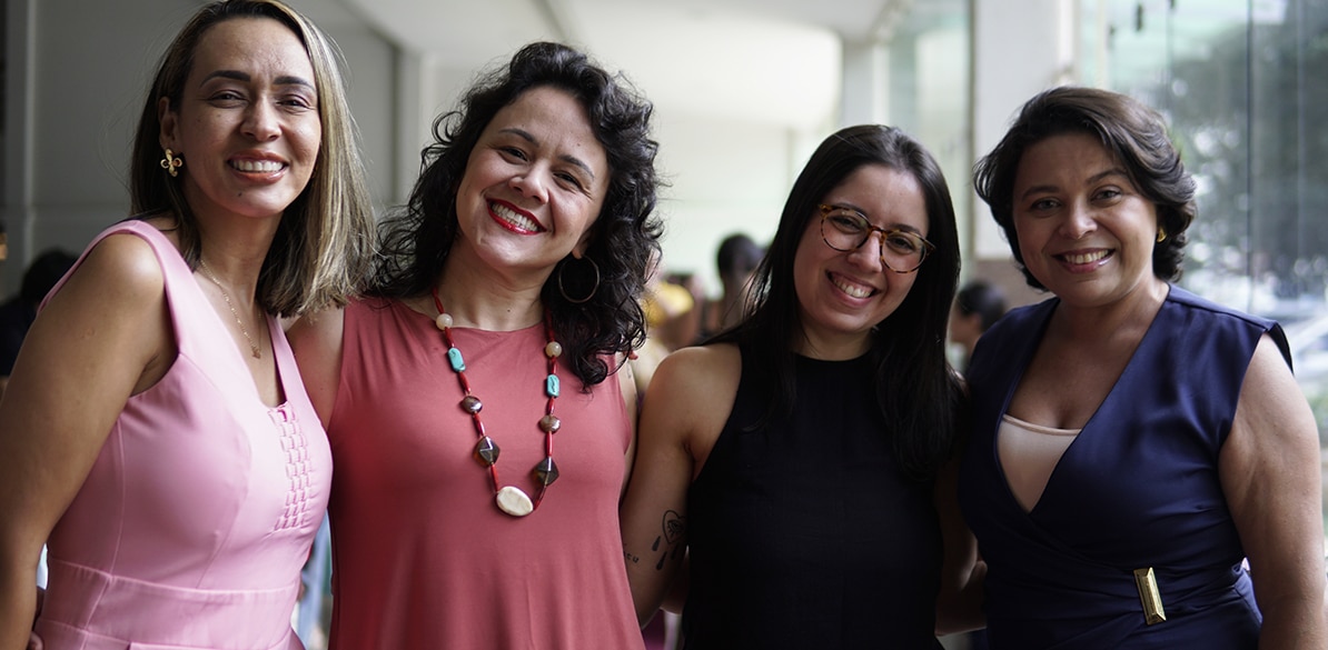 Educar também é cuidar Quatro profissionais da educação sorridentes e abraçadas posam juntas em um espaço iluminado durante um evento de reconhecimento ao trabalho docente.