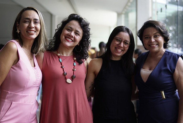 Educar também é cuidar Quatro profissionais da educação sorridentes e abraçadas posam juntas em um espaço iluminado durante um evento de reconhecimento ao trabalho docente.