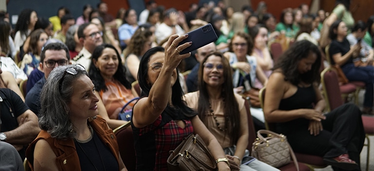 Educar também é cuidar Mulher fazendo uma selfie com o celular em meio ao público de um auditório lotado de docentes durante um evento sobre saúde mental nas escolas.