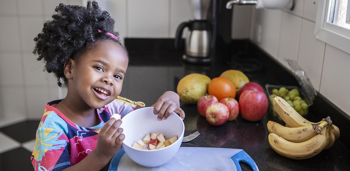 Dia Mundial da Saúde Menina negra pequena sorrindo enquanto come frutas picadas em uma tigela branca na cozinha, com bananas, maçãs, uvas e manga ao fundo, representando hábitos alimentares saudáveis desde a infância.