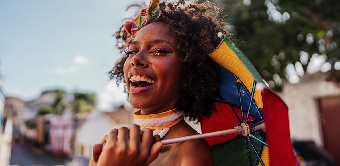 Mulher sorridente com maquiagem de Carnaval e adereço no cabelo, segurando sombrinha de frevo colorida em rua ao ar livre.