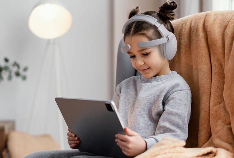 Menina com fones de ouvido brancos usando um tablet sentada em uma poltrona em casa, representando neurotecnologia pediátrica para crianças com TDAH e autismo.